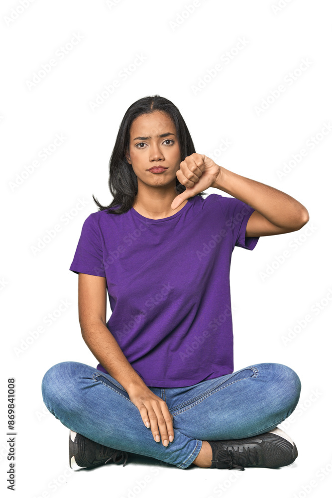 Latina sitting on floor in studio showing a dislike gesture, thumbs down. Disagreement concept. Latina sitting on floor in studio showing a dislike gesture, thumbs down. Disagreement concept.