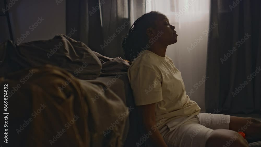 Side shot of young Black woman in loungewear sitting on floor by unmade bed and taking tranquilizing drug after sleepless night, living with insomnia Side shot of young Black woman in loungewear sitting on floor by unmade bed and taking tranquilizing drug after sleepless night, living with insomnia