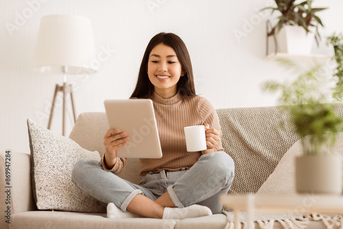 A young woman sits on a couch in a living room, smiling as she reads on her tablet while holding a cup of coffee.