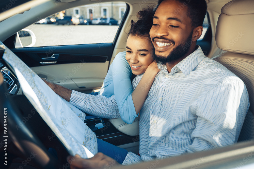 A young black couple sits in the front seats of a car, with the man driving and holding a map. The woman is leaning into the man with her arms wrapped around him.