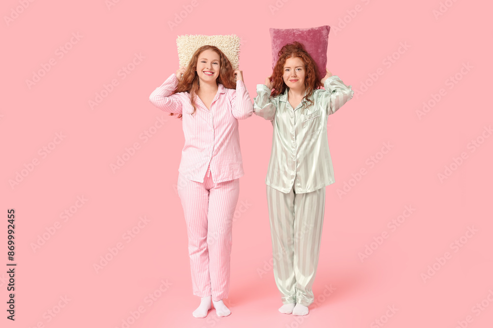 Happy redhead sisters in pajamas with pillows on pink background Happy redhead sisters in pajamas with pillows on pink background