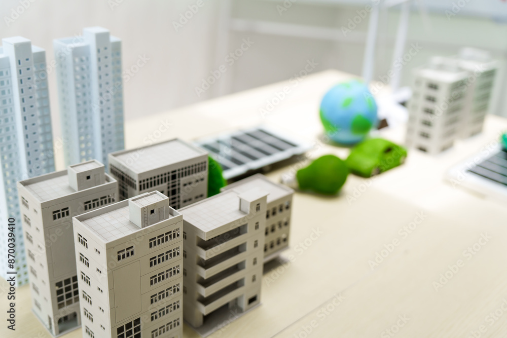 A white desk displays a model of an electric car, a green light bulb, and solar cells. The backdrop features city buildings and a wind turbine, symbolizing green energy and sustainability. A white desk displays a model of an electric car, a green light bulb, and solar cells. The backdrop features city buildings and a wind turbine, symbolizing green energy and sustainability.