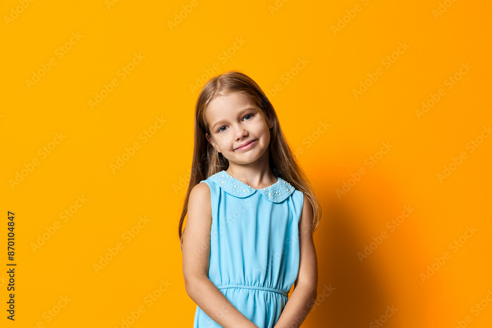 Little girl in a blue dress with hands in pockets stands confidently against a vibrant orange background Little girl in a blue dress with hands in pockets stands confidently against a vibrant orange background