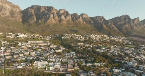 Drone flying high backwards on top of camps bay in cape town south africa - many houses on a slope - view of table mountain and the wide sea at sunset