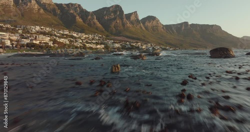 Drone flies just over the sea at camps bay in cape town south africa - wavy sea, many houses on a hilltop - drone flies with view to table mountain rocks in the sea