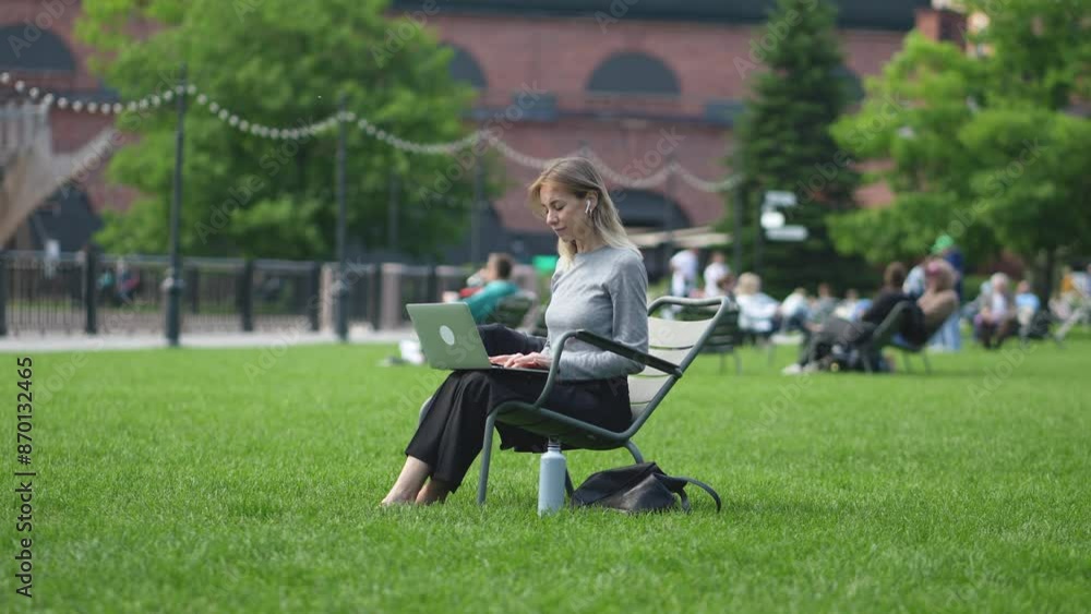 Focused woman with computer sitting on folding chair on lawn in busy public place. Peaceful businesswoman working in open air during day. Calm freelance girl attentively performing project tasks. Focused woman with computer sitting on folding chair on lawn in busy public place. Peaceful businesswoman working in open air during day. Calm freelance girl attentively performing project tasks.