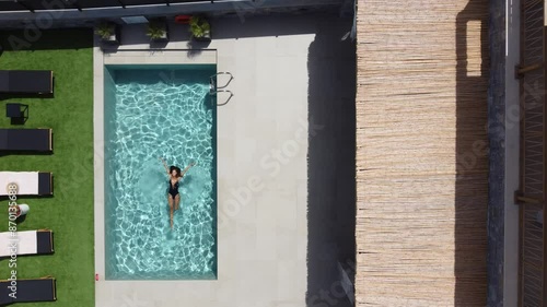 Drone flies over an outdoor pool in bird's eye view - Woman in swimming costume floats on her back on water - Villa in Greece Crete with palm trees photo