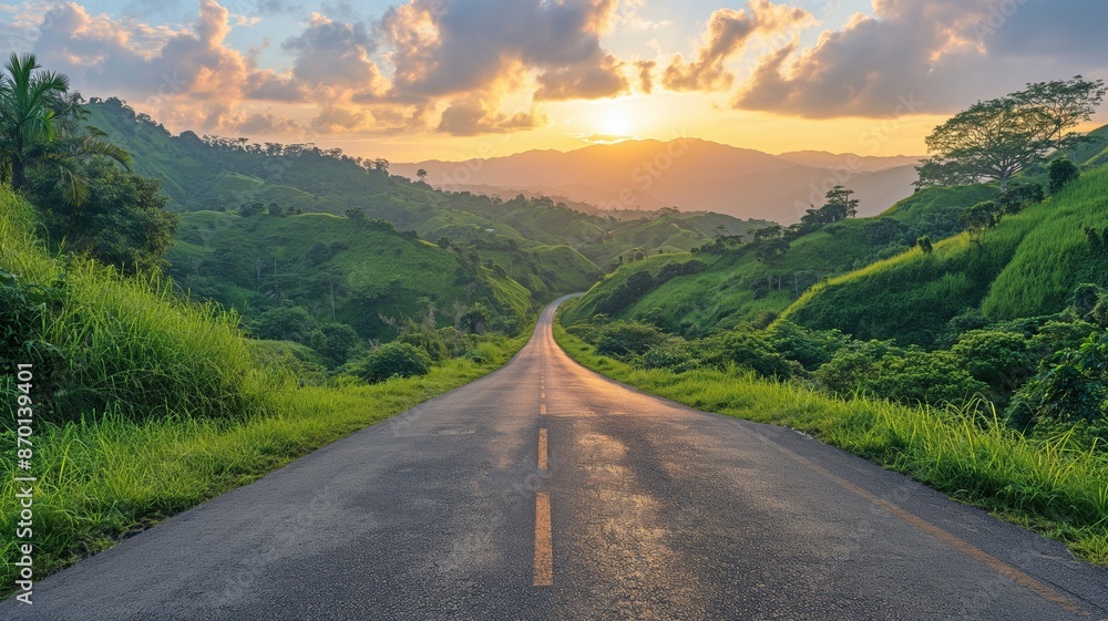 Naklejka premium A scenic road winds through lush, green hills as the sun rises, casting a warm glow over the landscape.