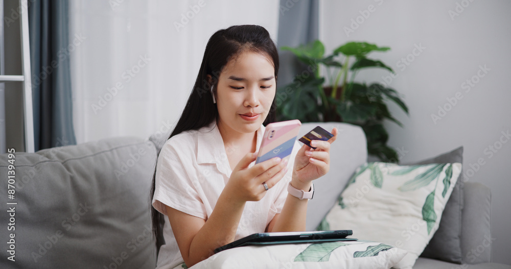 Portrait of Happy young woman sitting on sofa using credit card to pay on mobile phone in the living room at home ,Buy things online by phone, Pay online. Portrait of Happy young woman sitting on sofa using credit card to pay on mobile phone in the living room at home ,Buy things online by phone, Pay online.