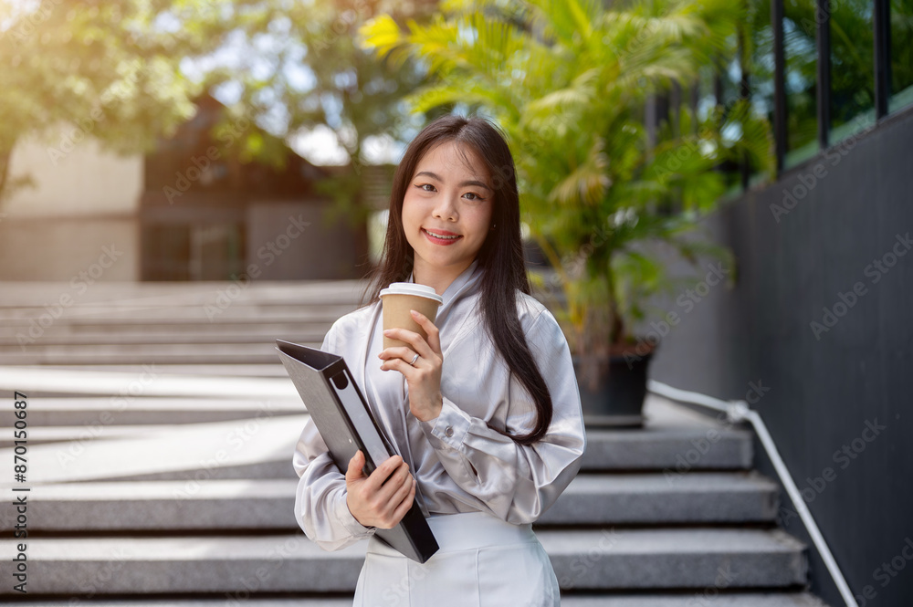 Obraz premium Attractive Asian businesswoman with a file folder and a coffee cup, standing on the stairs outdoors.