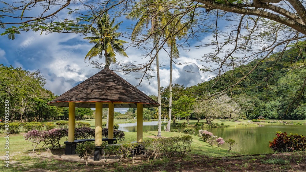 Landscape design of the park. A gazebo for relaxing next to the pond. Conical roof, columns, benches in the shade. Flowering bougainvilleas are nearby. Coconut palms against the blue sky and clouds Landscape design of the park. A gazebo for relaxing next to the pond. Conical roof, columns, benches in the shade. Flowering bougainvilleas are nearby. Coconut palms against the blue sky and clouds