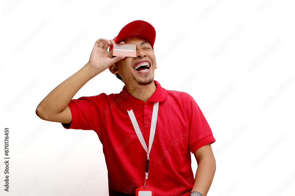 An excited Asian delivery man or courier playfully covers half of his face with a credit card, isolated on a white background. Concepts of online transactions, secure payments, customer satisfaction An excited Asian delivery man or courier playfully covers half of his face with a credit card, isolated on a white background. Concepts of online transactions, secure payments, customer satisfaction