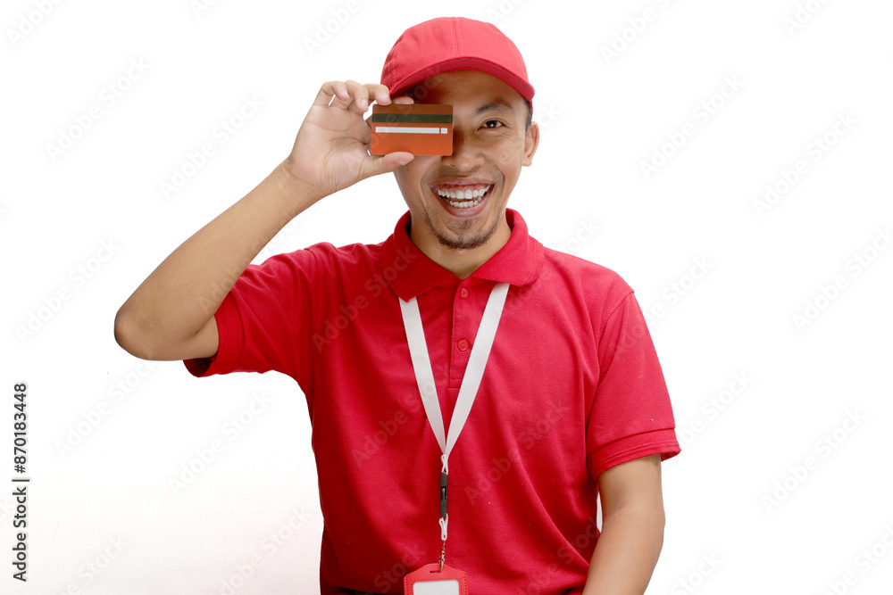 An excited Asian delivery man or courier playfully covers half of his face with a credit card, isolated on a white background. Concepts of online transactions, secure payments, customer satisfaction An excited Asian delivery man or courier playfully covers half of his face with a credit card, isolated on a white background. Concepts of online transactions, secure payments, customer satisfaction