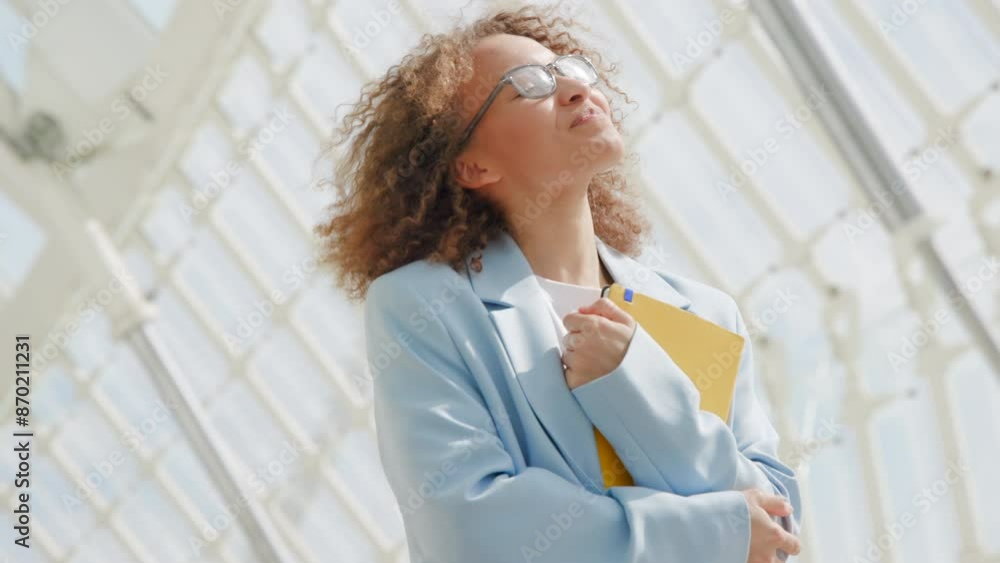 Within a modern atrium, a young businesswoman with curly hair exudes confidence in a light blue blazer, holding a yellow folder. The elegant workplace reflects professionalism and a bright environment Within a modern atrium, a young businesswoman with curly hair exudes confidence in a light blue blazer, holding a yellow folder. The elegant workplace reflects professionalism and a bright environment