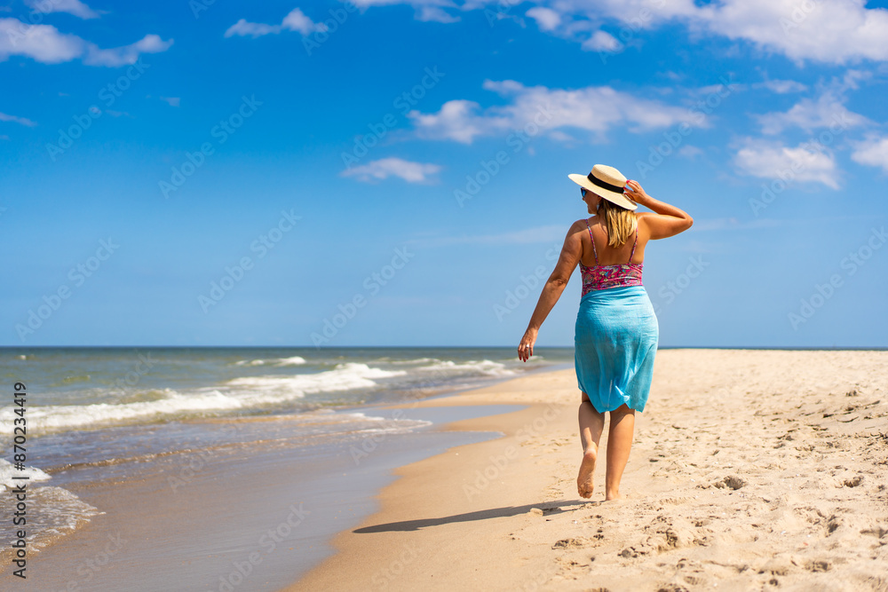 Summer vacation. Beautiful young woman in colorful swimsuit and blue pareo and hat walking on the sandy beach on a beautiful sunny day. Back view Summer vacation. Beautiful young woman in colorful swimsuit and blue pareo and hat walking on the sandy beach on a beautiful sunny day. Back view