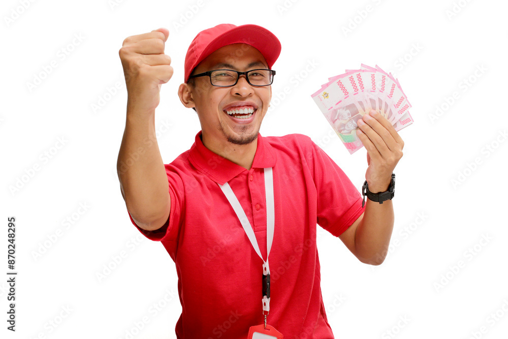 Excited Asian delivery man or courier raises his fist in triumphant YES gesture while holding money banknotes in his hand, isolated on white background. Concepts of victory, financial gains, triumphs Excited Asian delivery man or courier raises his fist in triumphant YES gesture while holding money banknotes in his hand, isolated on white background. Concepts of victory, financial gains, triumphs