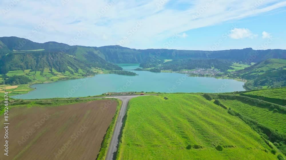 Lush green fields, a winding road, and a serene blue lake surrounded by mountains, aerial view Lush green fields, a winding road, and a serene blue lake surrounded by mountains, aerial view