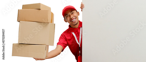 Excited Asian delivery man or courier peeks out from behind a white wall with copy space, holding a cardboard box, isolated on a white background. Delivery services, shipping, or e-commerce businesses photo