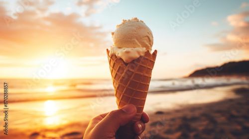 A hand holds a single-scoop ice cream cone in a waffle cone on a beach at sunset © reddish