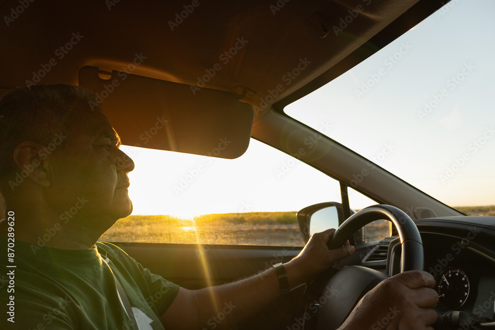 Naklejka premium Adult man driving a car during golden hour, holds the steering wheel with confidence and security on a road trip on vacation during sunset