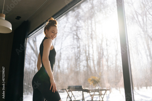 Low angle back view of smiling fitness female posing at home standing by window with beautiful snowy winter nature view on sunny day. Attractive successful woman coach in sportswear with closed eyes. © dikushin