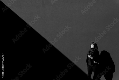 Woman with a Smartphone Standing Against a Grey Wall with Diagonal Shadow in a Sunny Day in Switzerland.