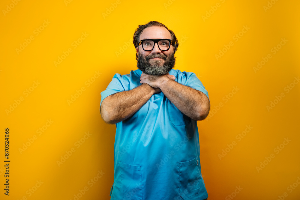 The image shows a man with a beard, glasses and a blue shirt standing in front of a yellow background. He is looking up, with his mouth open and his hands clasped around his neck. The image shows a man with a beard, glasses and a blue shirt standing in front of a yellow background. He is looking up, with his mouth open and his hands clasped around his neck.