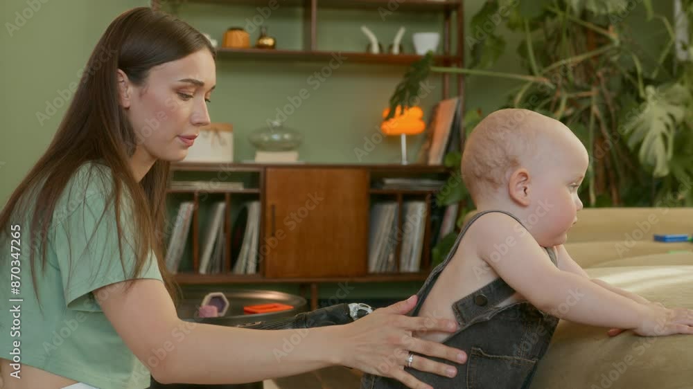 Young mother with prosthetic changes clothes her little son sitting on floor during playing toys in living room. Young woman with prosthesis hand takes care of her baby boy Young mother with prosthetic changes clothes her little son sitting on floor during playing toys in living room. Young woman with prosthesis hand takes care of her baby boy
