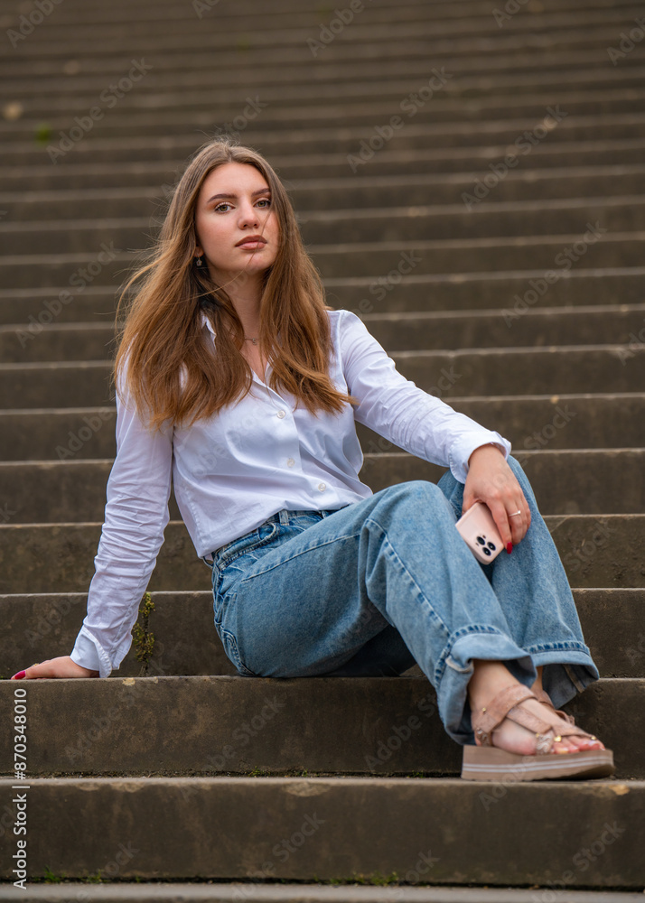 A modern young girl with long flowing hair sits on stone steps in the city. Looking at the camera, holding a mobile phone in his hands. A modern young girl with long flowing hair sits on stone steps in the city. Looking at the camera, holding a mobile phone in his hands.