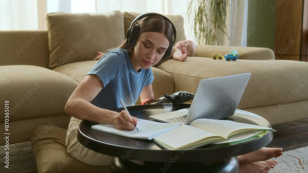 Young mother with prosthetic arm works remotely on laptop while baby naps on couch in living room. Female student in headphones sits on floor and studies for exam using notebooks and online resources. Young mother with prosthetic arm works remotely on laptop while baby naps on couch in living room. Female student in headphones sits on floor and studies for exam using notebooks and online resources.
