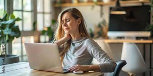 A woman is sitting at a desk with a laptop and a mouse. She is wearing a gray shirt and she is in pain