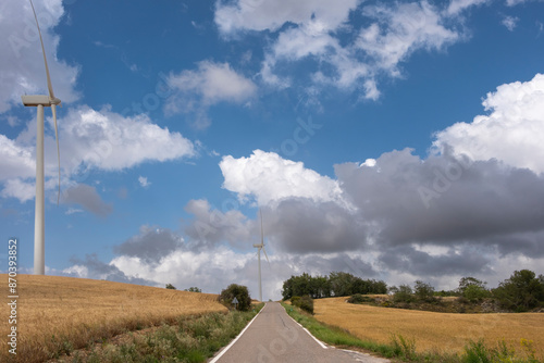 A deserted rural road stretches ahead with tall wind turbines and golden fields on either side, under a partly cloudy blue sky, evoking peace and sustainability. photo