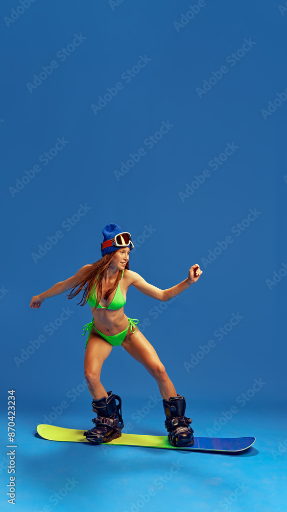 Obraz premium Stylish woman in bikini and winter sports gear, demonstrating her adventurous spirit and love for snowboarding against blue studio background. Concept of vacation, tourism, active lifestyle, fashion