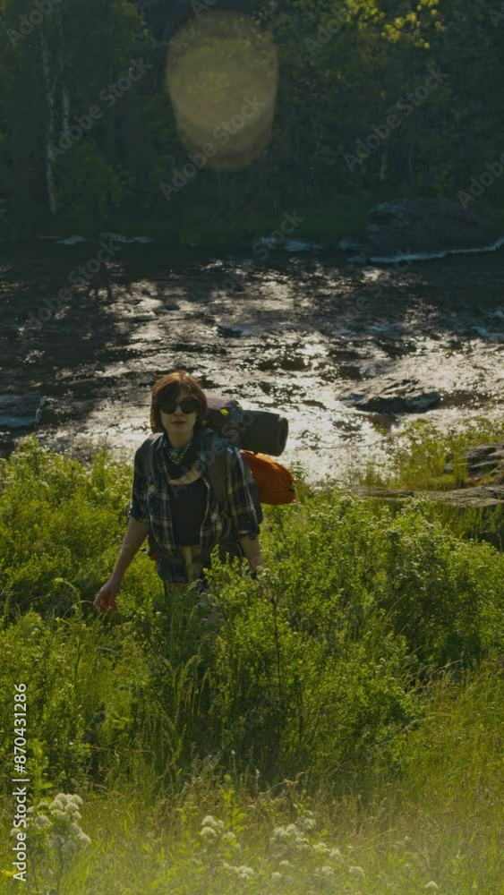 Vertical full shot of young woman in sunglasses hiking on sunlit green grass beside river in summer landscape Vertical full shot of young woman in sunglasses hiking on sunlit green grass beside river in summer landscape