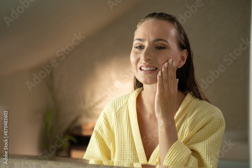 A woman stands in the bathroom, looking in the mirror while experiencing tooth pain. She is touching her jaw and face, grimacing in discomfort photo