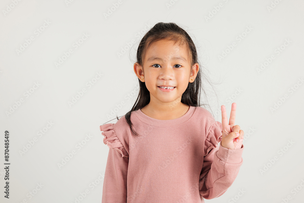 Asian young kid girl showcases optimism and peace by gesturing victory and peace signs in a studio shot with a white background. kindergarten child joyful smile speaks of success, V sign finger Asian young kid girl showcases optimism and peace by gesturing victory and peace signs in a studio shot with a white background. kindergarten child joyful smile speaks of success, V sign finger