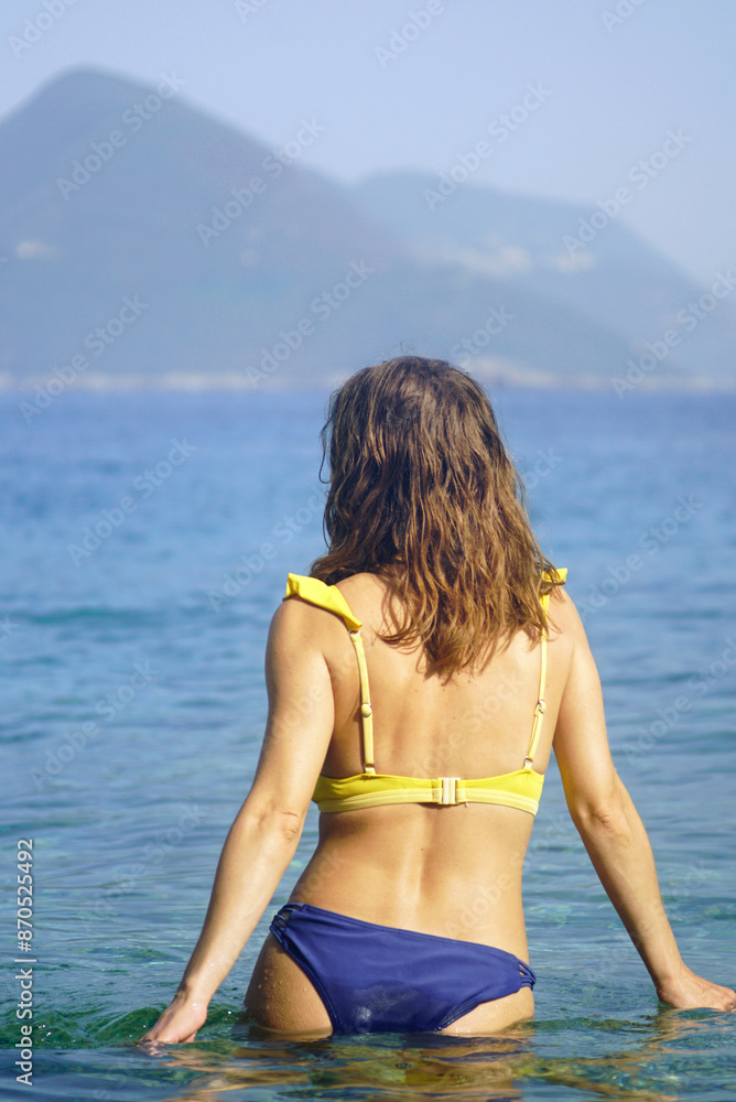 A girl with wavy hair, dressed in a bright two-piece swimsuit, walks into the sea in a picturesque place. A woman photographed from the back stands in the water against the backdrop of mountains A girl with wavy hair, dressed in a bright two-piece swimsuit, walks into the sea in a picturesque place. A woman photographed from the back stands in the water against the backdrop of mountains
