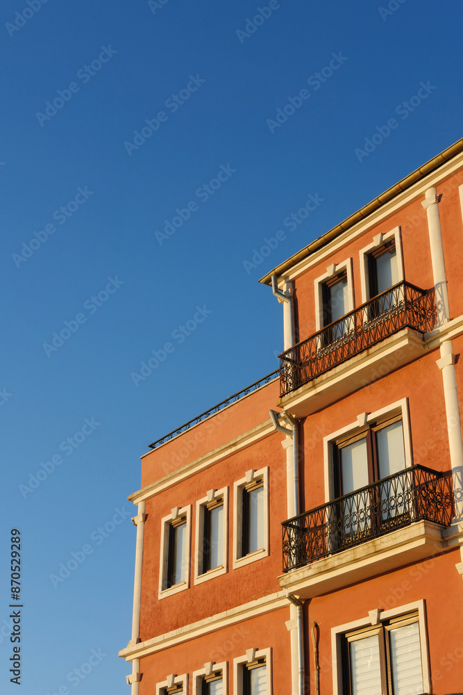 Sunlit Orange Building with Balconies at Dusk. An orange building with balconies illuminated by the warm light of the setting sun, contrasted against a clear blue sky. Highlights architectural details Sunlit Orange Building with Balconies at Dusk. An orange building with balconies illuminated by the warm light of the setting sun, contrasted against a clear blue sky. Highlights architectural details