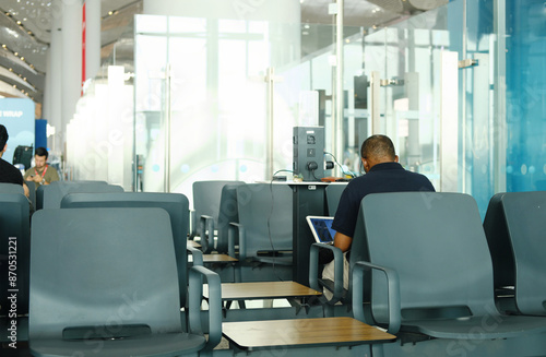 Man Working on Laptop in Airport Lounge. A man sitting in an airport lounge using a laptop, surrounded by empty gray chairs and modern decor.