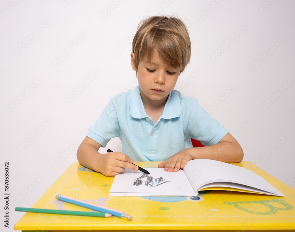 Smiling cute small european boy sitting at table, draws picture on withe background. Smiling cute small european boy sitting at table, draws picture on withe background.