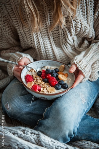 Healthy winter breakfast in bed. A woman in a wool sweater and faded jeans eats vegan almond milk oatmeal in a bowl with berries, fruits and almonds. 