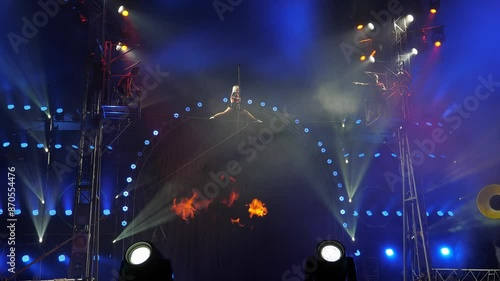 A circus show with tightrope walkers under the dome of the circus under the light of colorful spotlights. An acrobat girl sits on a rope splits under the dome of the circus.