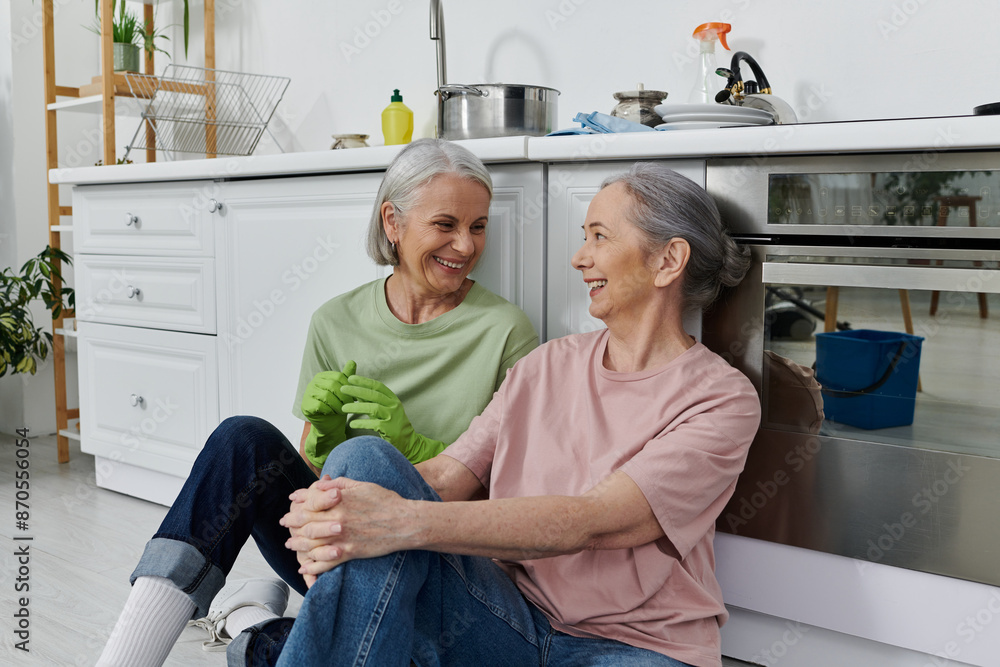 A mature lesbian couple, dressed casually, are cleaning their modern apartment together, taking a break to share a laugh. A mature lesbian couple, dressed casually, are cleaning their modern apartment together, taking a break to share a laugh.