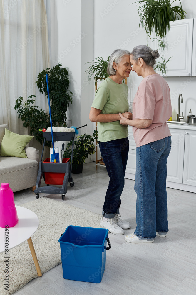 Two mature women, a lesbian couple, take a break from cleaning their modern apartment. They are standing in the living room, holding hands. Two mature women, a lesbian couple, take a break from cleaning their modern apartment. They are standing in the living room, holding hands.