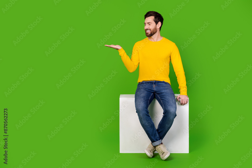 Full length photo of handsome young guy hold look empty space sit cube dressed stylish yellow garment isolated on green color background Full length photo of handsome young guy hold look empty space sit cube dressed stylish yellow garment isolated on green color background