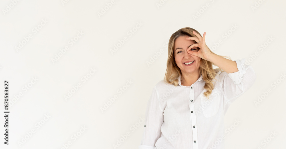 Happy young woman shows ok sign with hand on eye looking through fingers on white background. Happy young woman shows ok sign with hand on eye looking through fingers on white background.