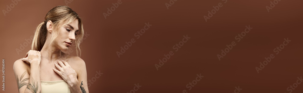 Fototapeta premium A young woman with vitiligo and a tattoo poses with her shoulders bare against a beige background.
