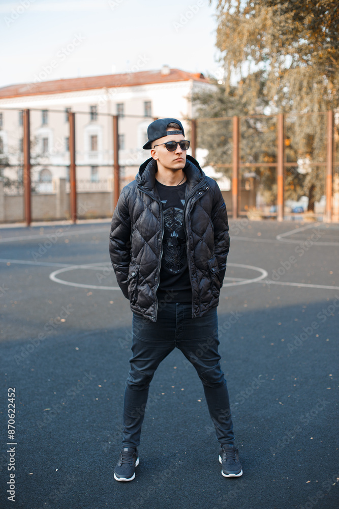 Cool fashionable stylish hipster guy with a baseball cap with sunglasses in a fashion down jacket with jeans and sneakers on the street at the stadium. Fashion male style outwear Cool fashionable stylish hipster guy with a baseball cap with sunglasses in a fashion down jacket with jeans and sneakers on the street at the stadium. Fashion male style outwear