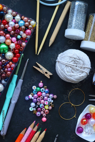 Colorful beads, coloring pencils, glitter, needles, string, hoop, tape and scissors on dark background. Various colorful craft supplies. Flat lay.