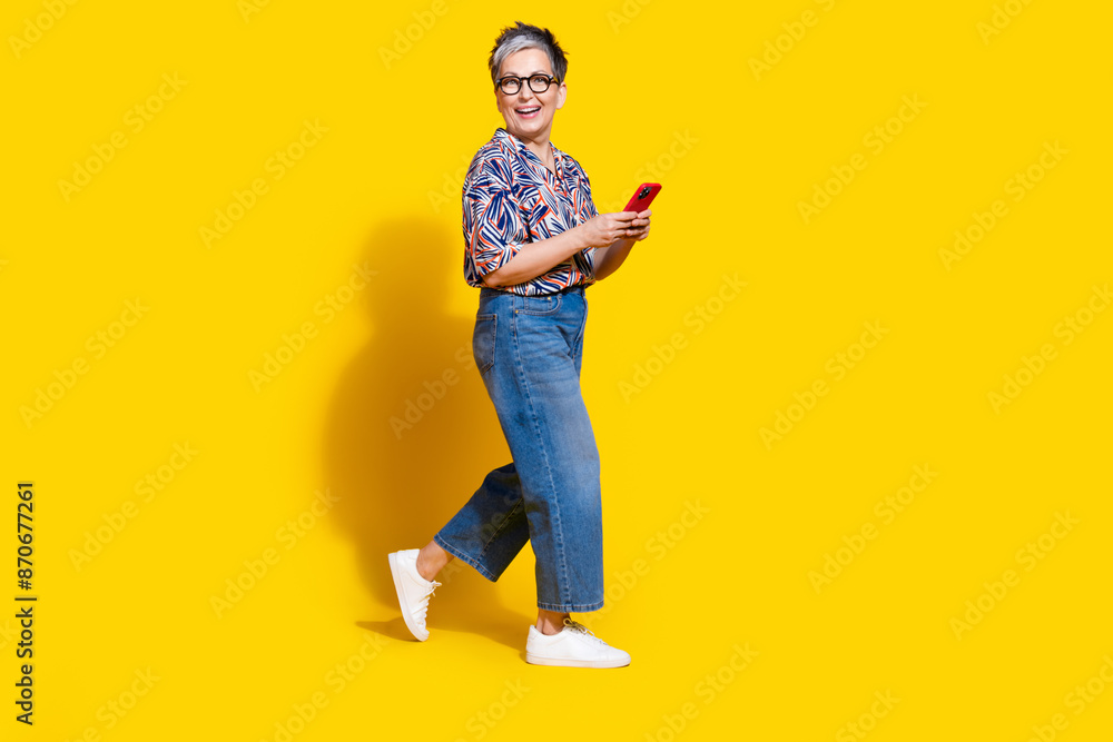Fototapeta premium Full body photo of attractive retired woman walk hold telephone dressed stylish colorful clothes isolated on yellow color background